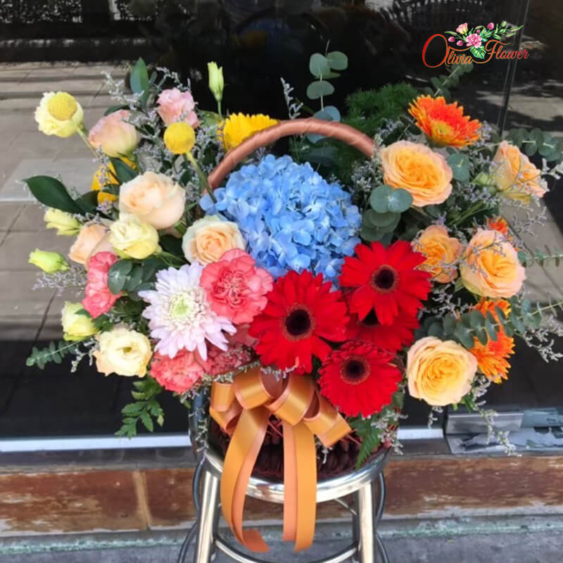 กระเช้าดอกไม้สด ประกอบด้วย ไฮเดรนเยียสีฟ้า กุหลาบสีส้ม เยอบีร่า สีแดง สีเหลือง สีส้ม คาร์เนชั่นสีส้ม และไลเซนทรัส